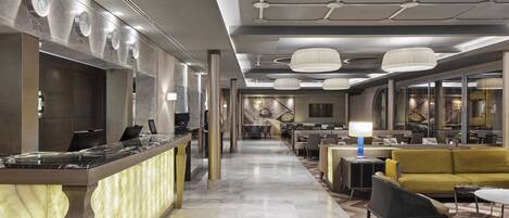 A modern hotel lobby with a reception desk, seating area, and a staircase leading to upper floors.