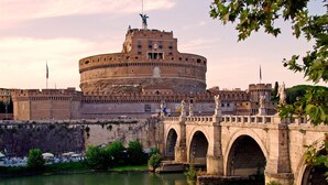 A historic fortress with a central round tower, a bridge with arches, and statues.