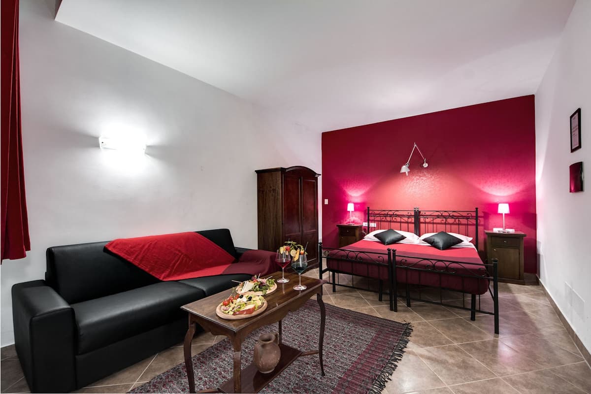 Quarto com uma cama, sofá preto, uma mesinha com comida e bebidas, e uma parede de destaque vermelha.
