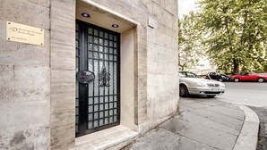 A building entrance with a metal gate, a plaque with a name and phone number, and a street with parked cars.