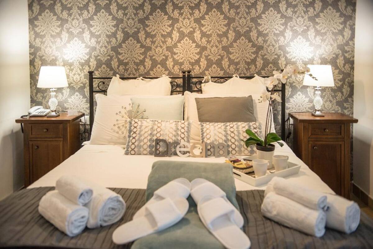 Cama bem arrumada com almofadas, mesa de cabeceira com abajur, bandeja com uma planta e toalhas dobradas na cama.