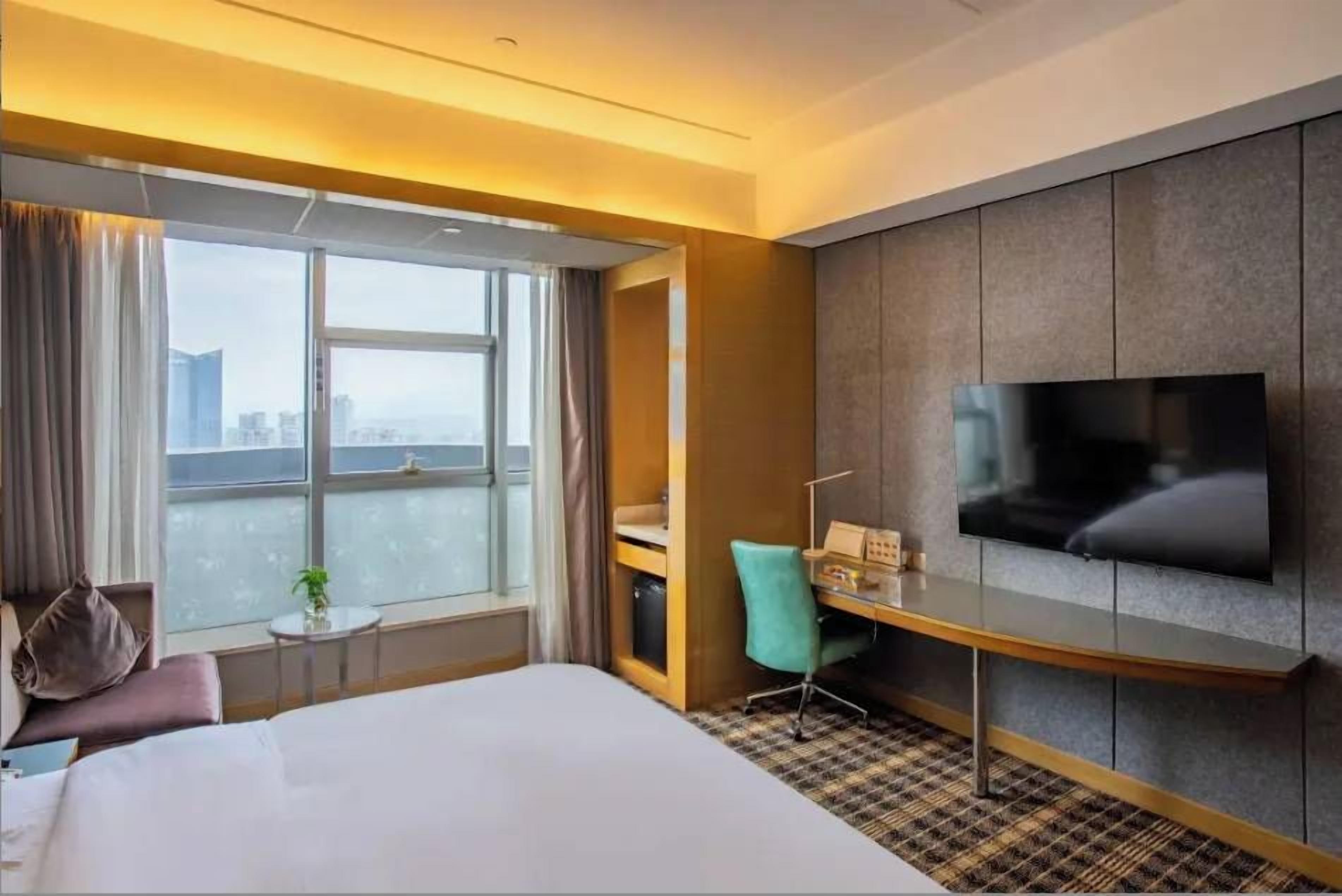 A hotel room with a bed, a desk, a chair, a television, and a window with a view of the city.
