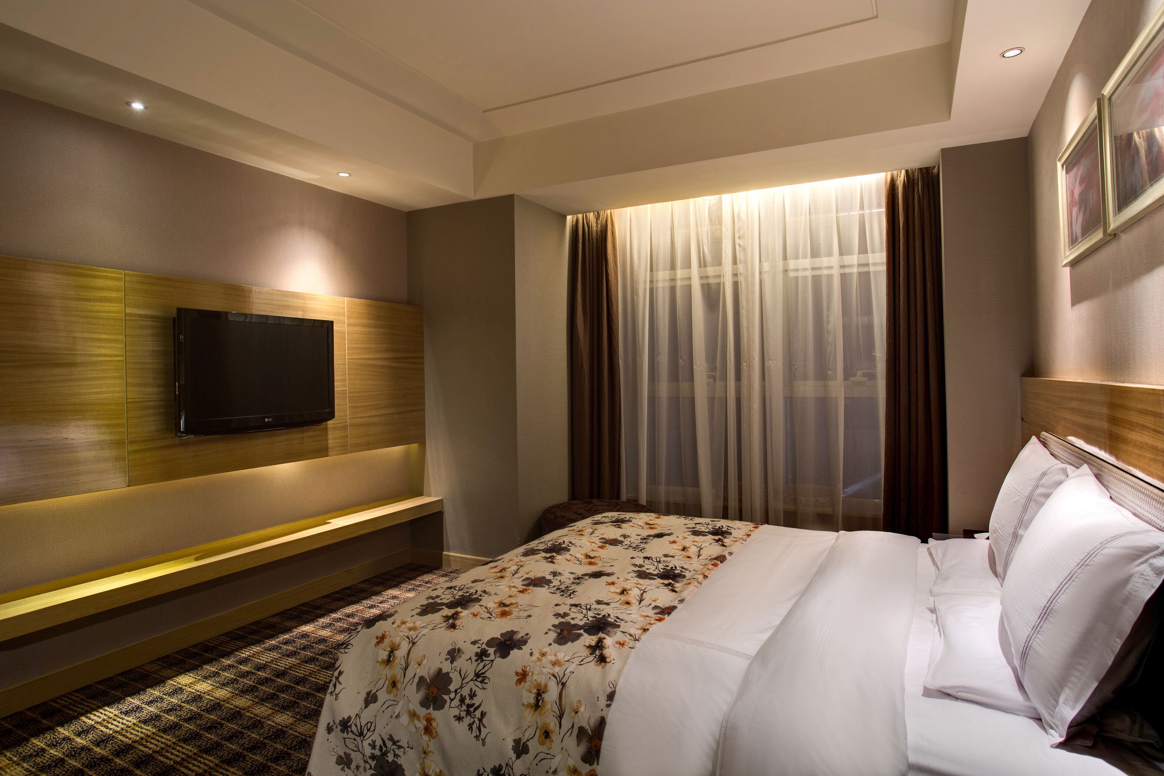 Una habitación de hotel con una cama grande, un televisor de pantalla plana montado en la pared, un banco y una ventana con cortinas traslúcidas.