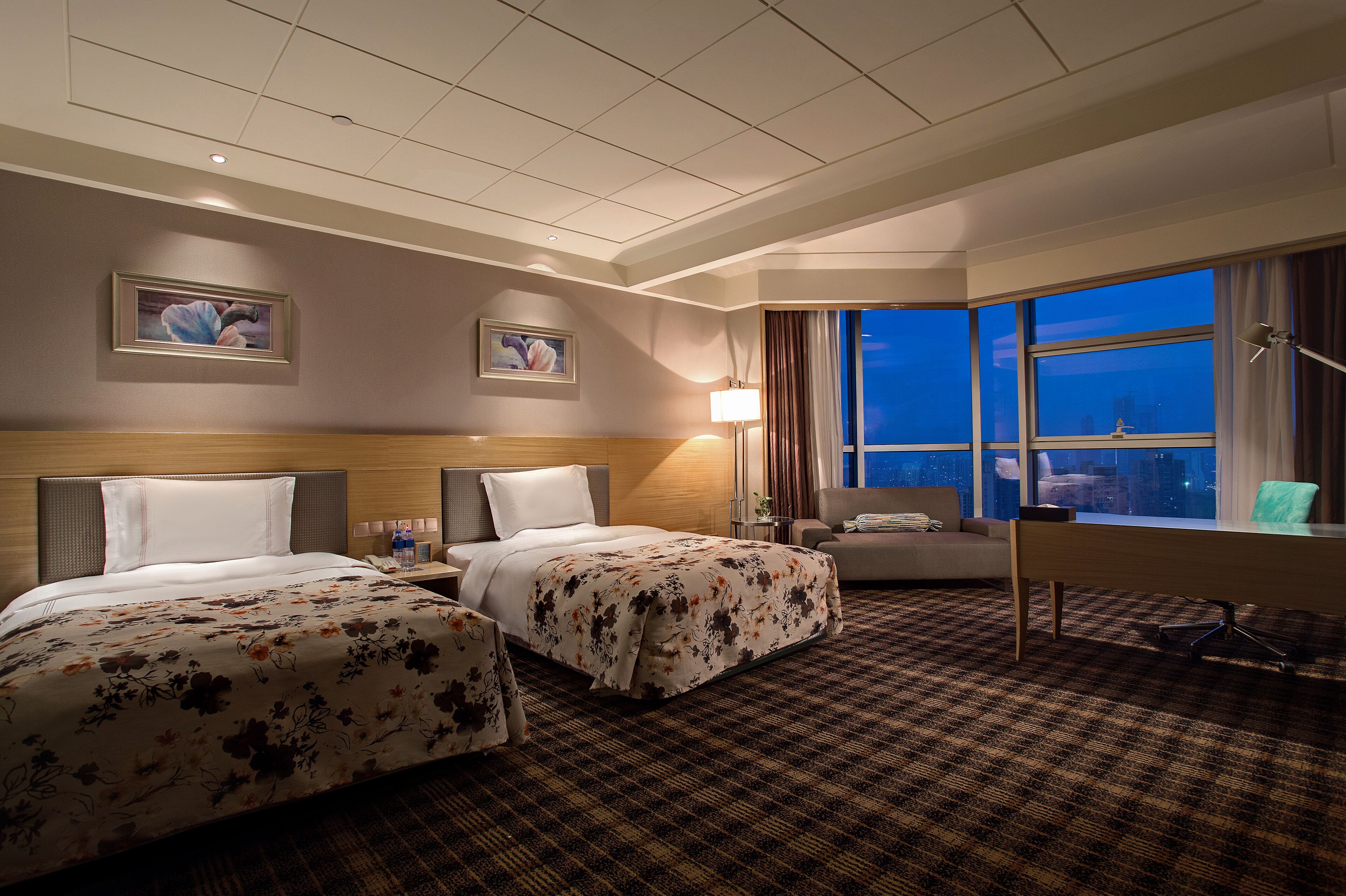 Ein Hotelzimmer mit zwei Betten, einem Schreibtisch, einem Stuhl, einer Lampe und einem großen Fenster mit Blick auf die Stadt.