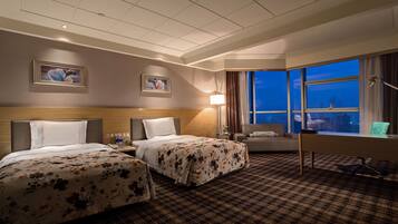 Ein Hotelzimmer mit zwei Betten, einem Schreibtisch, einem Stuhl, einer Lampe und einem großen Fenster mit Blick auf die Stadt.