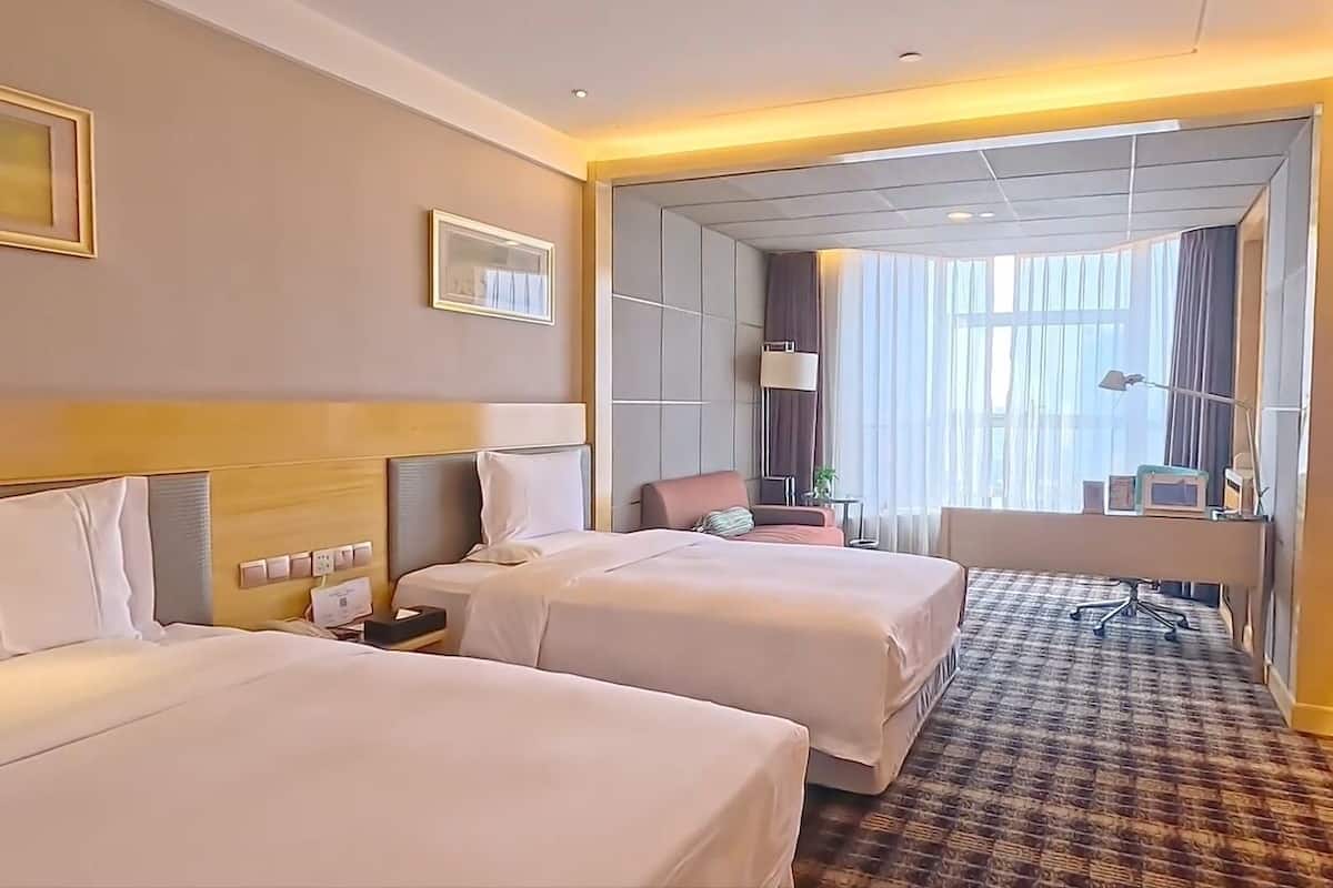 Une chambre d’hôtel avec deux lits, un bureau, une chaise, une lampe et une grande fenêtre avec des rideaux.