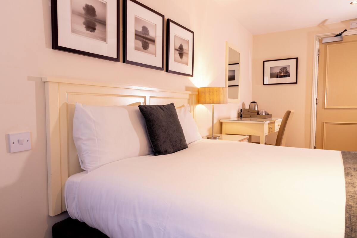 A hotel room with a bed, a desk, a chair, a lamp, and framed pictures on the wall.
