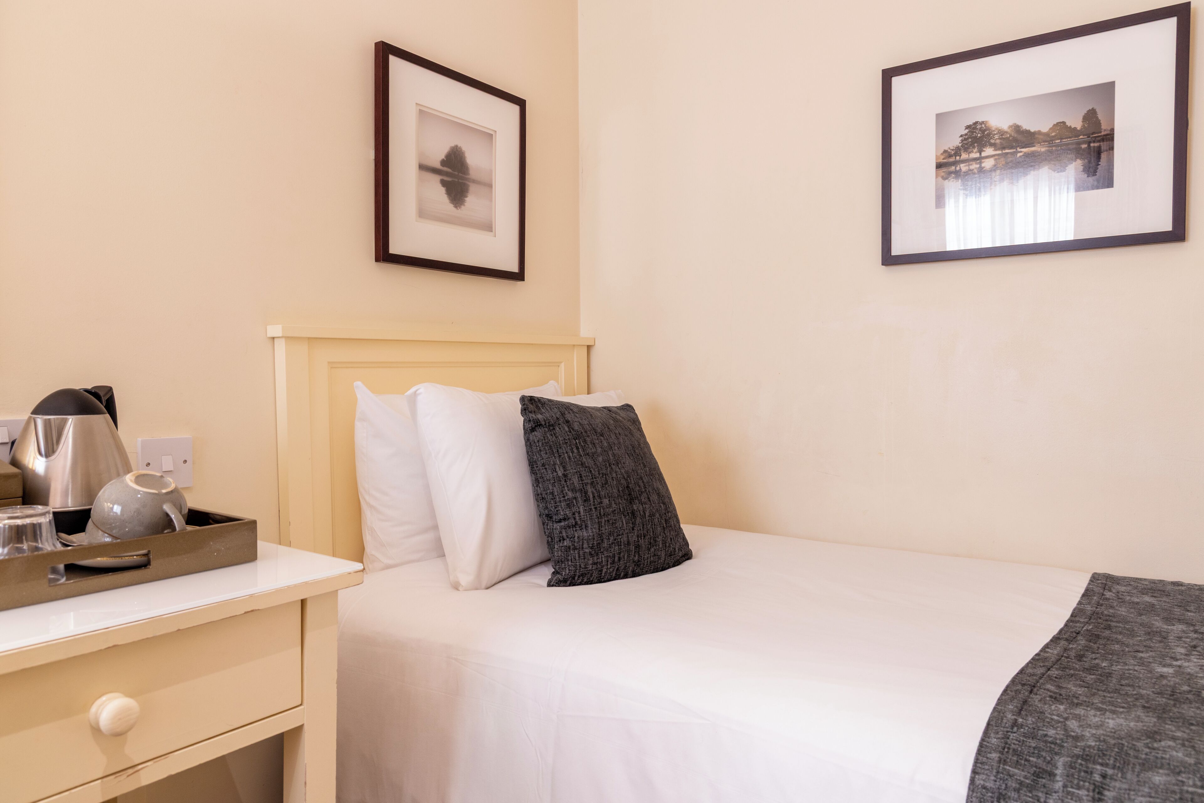 Habitación de hotel con una cama, una mesita de noche con una tetera y vasos, un cuadro en la pared y otro cuadro sobre la cama.