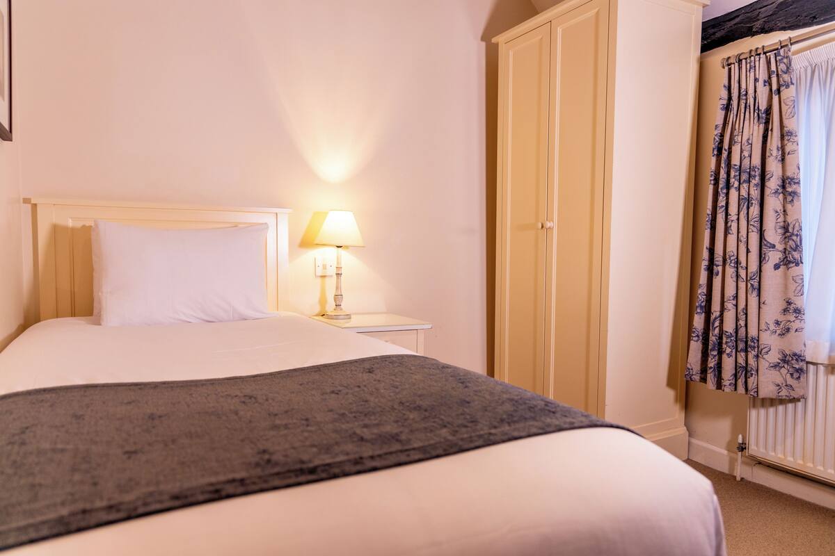 A bedroom with a bed, nightstand, lamp, wardrobe, and floral curtains.