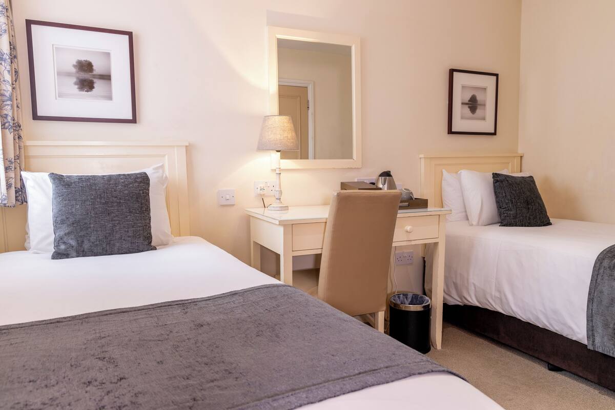 A hotel room with two beds, a desk, a chair, a lamp, a trash bin, and framed pictures on the walls.