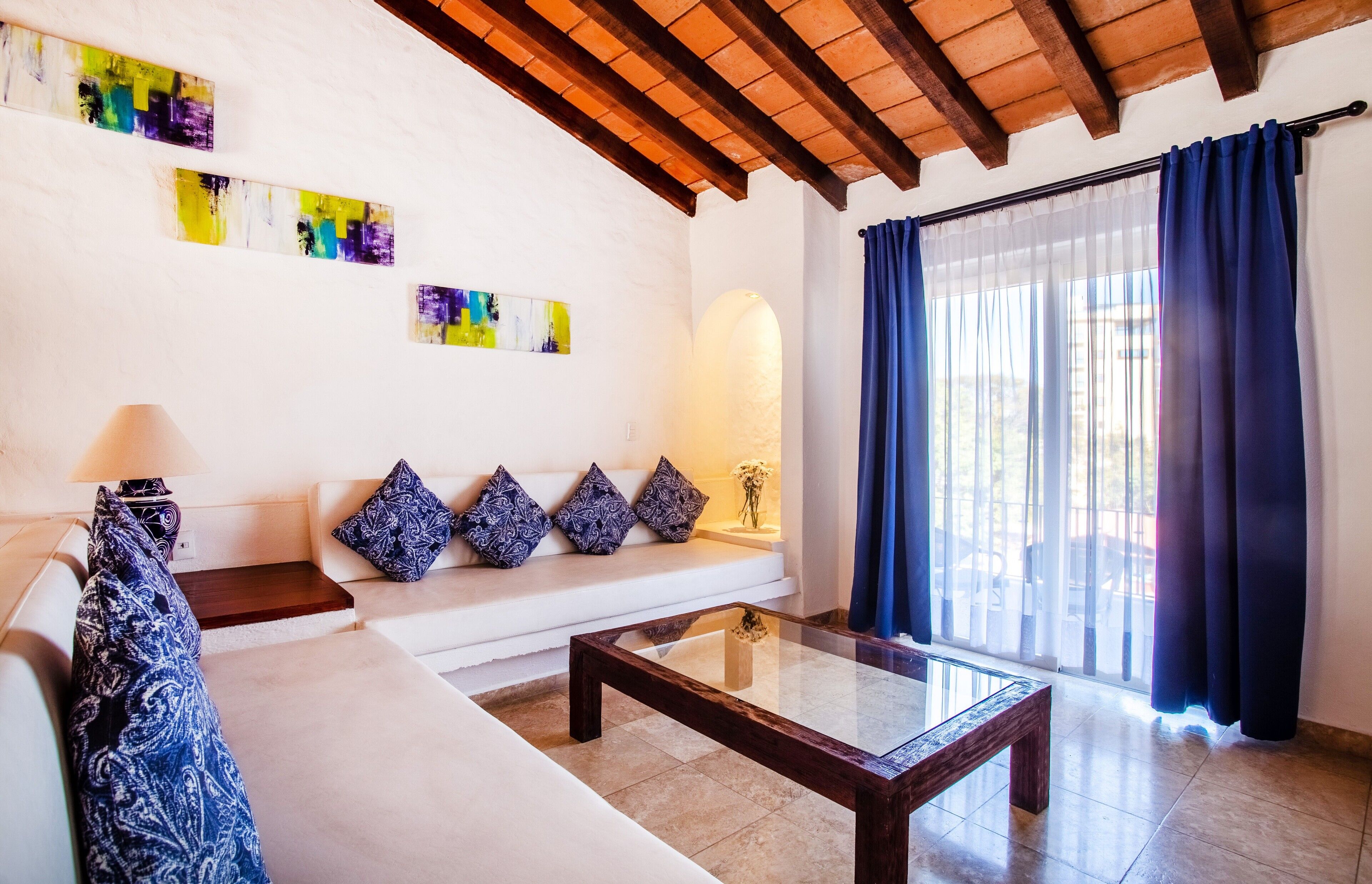 A living room with a white sofa, patterned cushions, a glass coffee table, and abstract wall art.