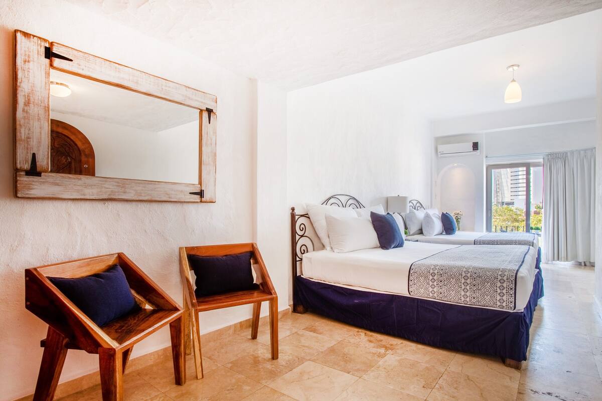 A bedroom with two wooden chairs, a bed with patterned bedding, a large mirror, and a view of the outdoors.
