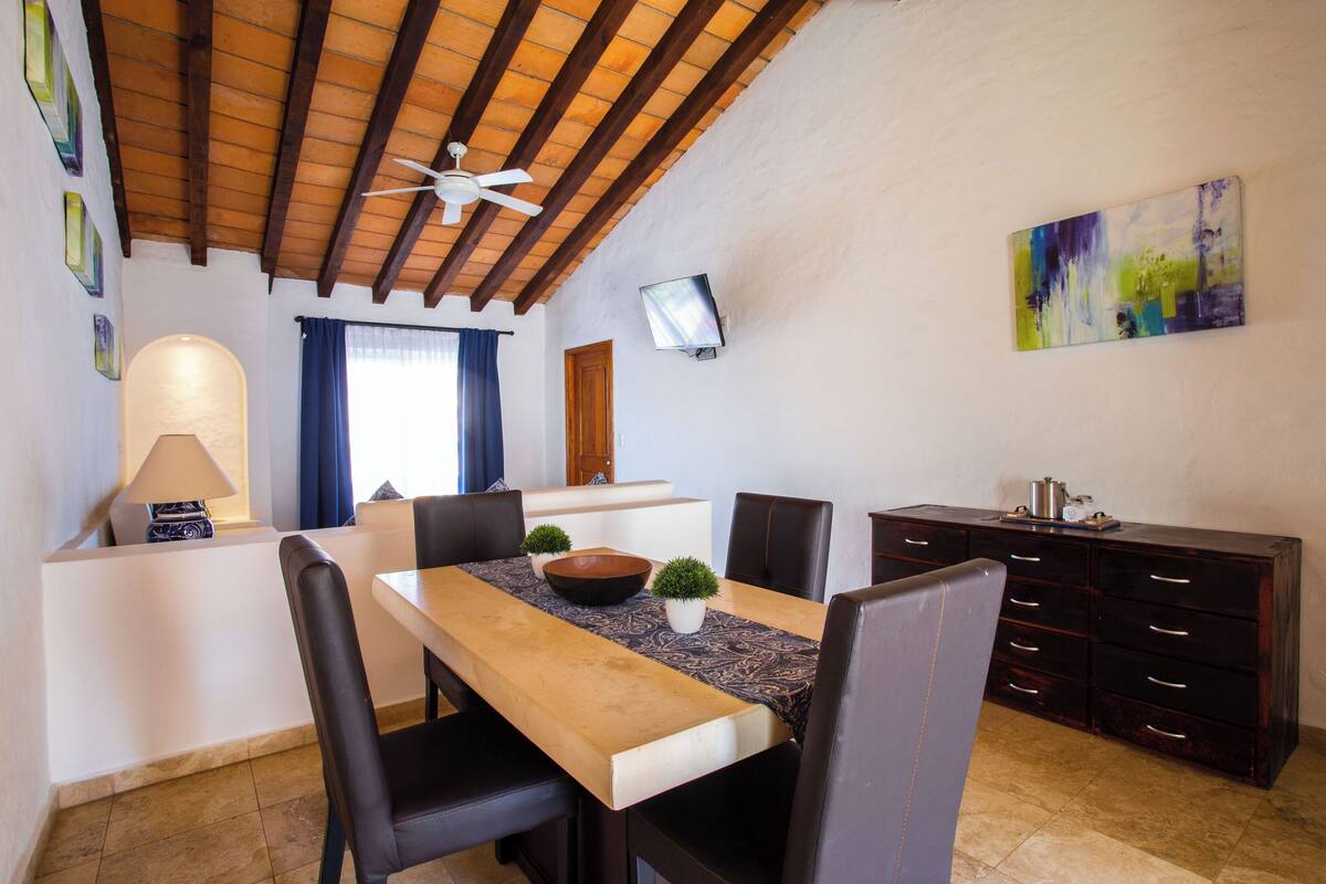 A dining area with a wooden table, black chairs, a ceiling fan, a TV on the wall, and a painting.