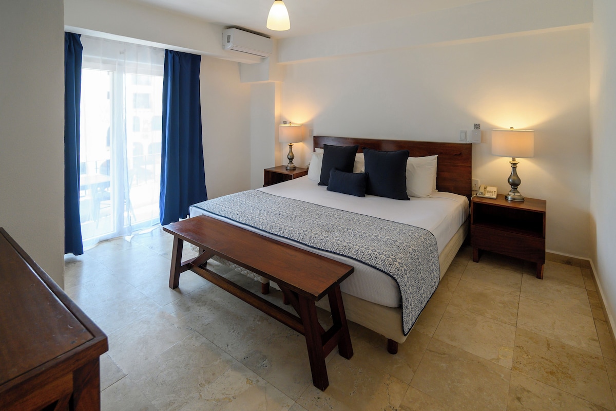 Habitación de hotel con cama, mesitas de noche, un banco y vistas al exterior.