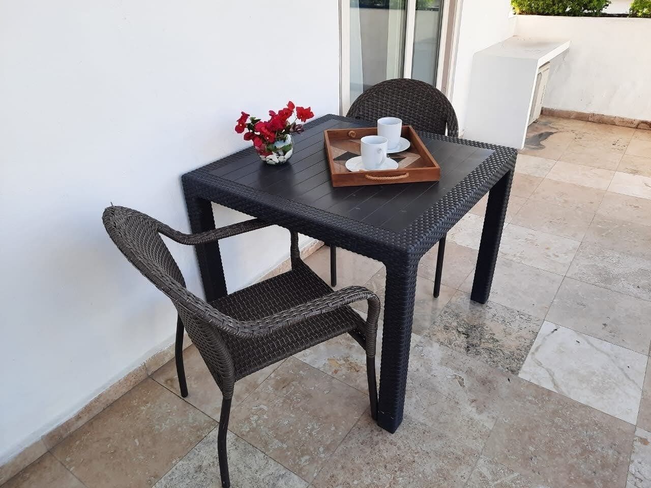 A small outdoor table with two wicker chairs, a tray with cups, and a vase with red flowers.