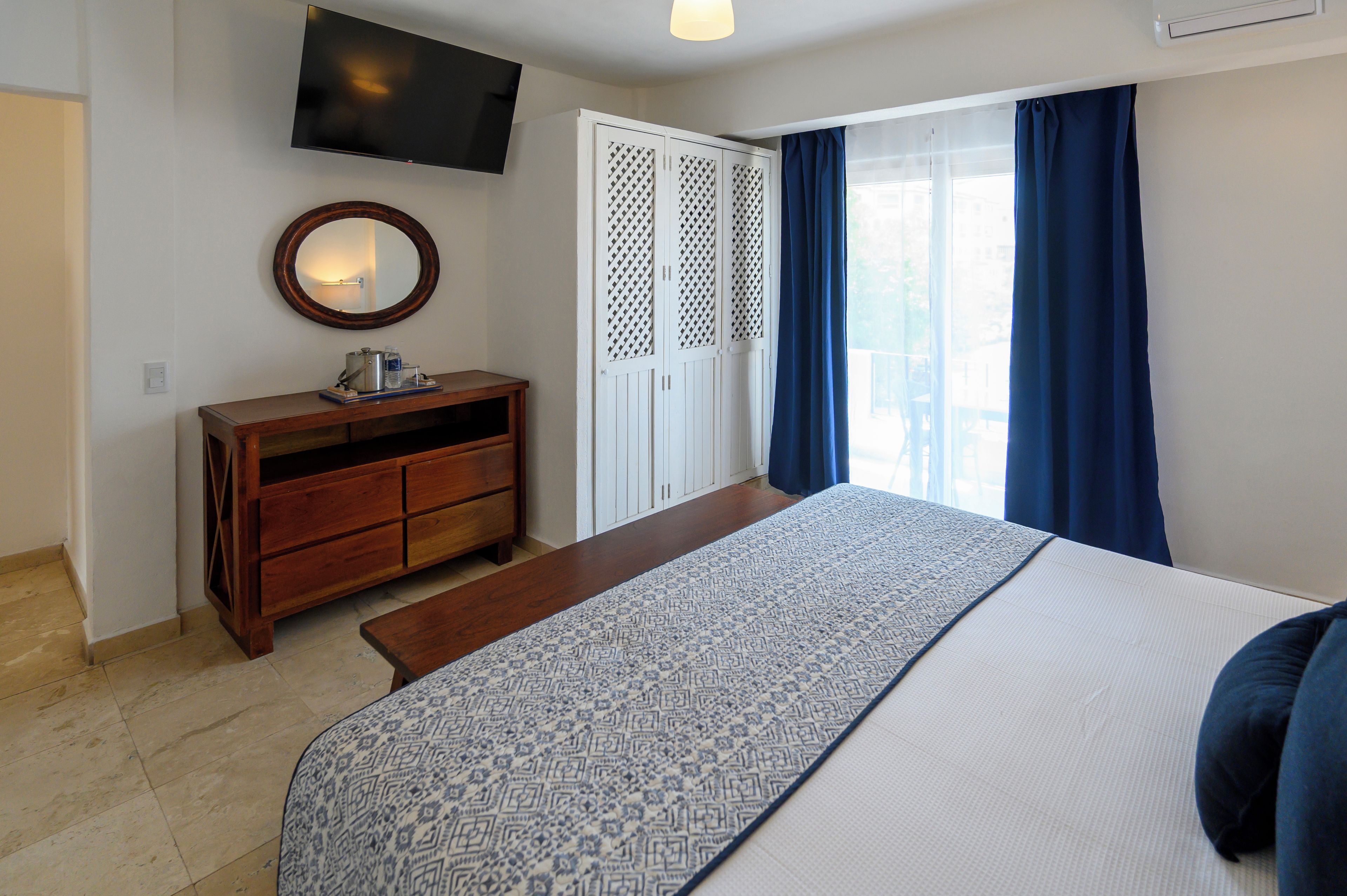 A hotel room with a bed, a wooden dresser, a television mounted on the wall, and a window with curtains.