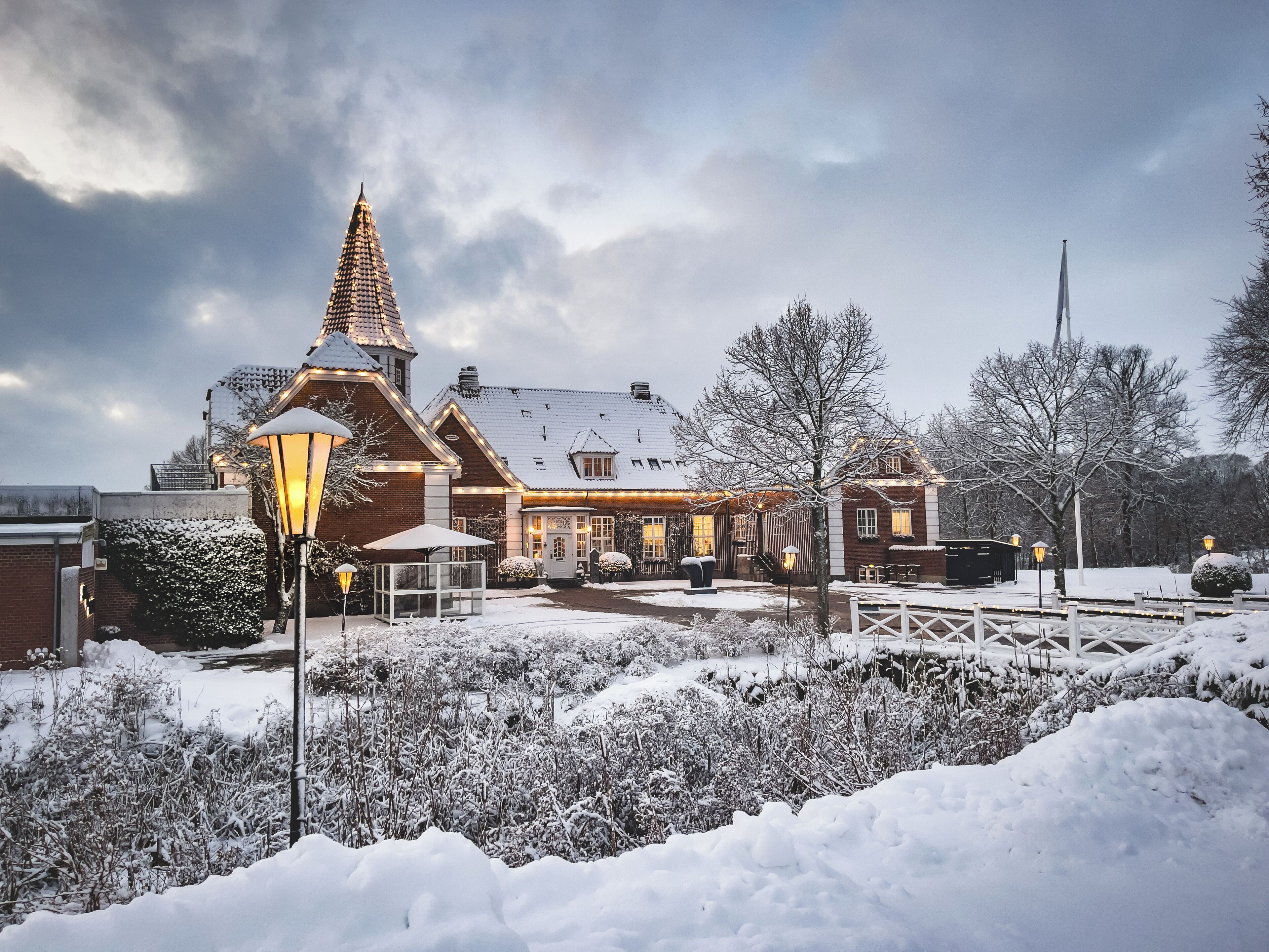 Un edificio cubierto de nieve con una torre destacada, un porche techado y un jardín nevado.