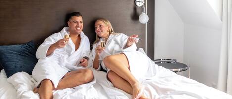 Una pareja vestida con túnicas blancas sobre una cama con cabecera de madera, una mesita de noche y una lámpara fijada a la pared.