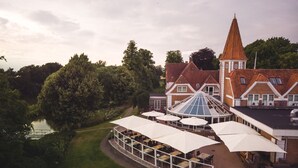 A riverside hotel with a dining area featuring outdoor umbrellas and tables, a large glass conservatory, and a traditional building with a conical roof. - Hotel Sørup Herregaard (Ringsted)