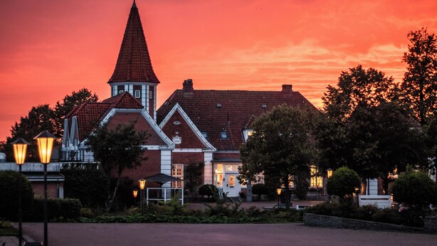 A building with a prominent tower and a red-tiled roof, surrounded by trees and street lamps. - Hotel Sørup Herregaard (Ringsted)