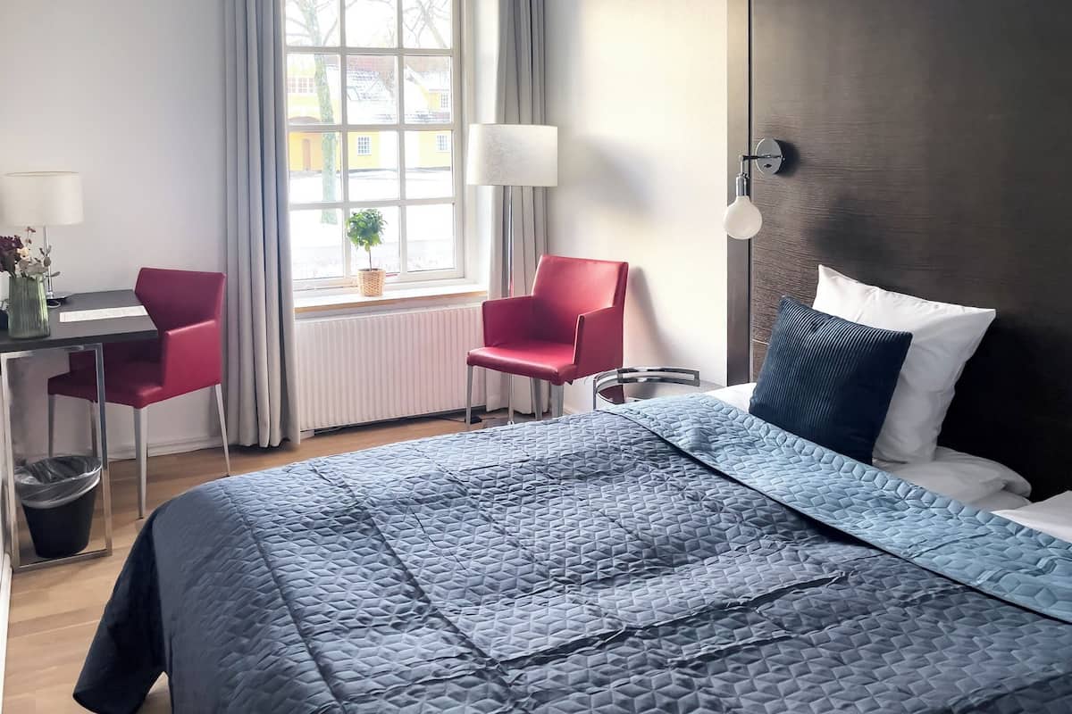 Ein Hotelzimmer mit einem großen Bett, einem Schreibtisch mit Stuhl, einem Fenster mit Vorhängen und einer Nachttischlampe.