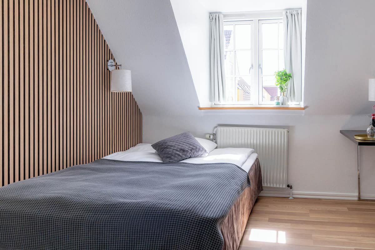 Ein Schlafzimmer mit schrägen Wänden, einem fenster mit Vorhängen, einer Heizung und einem Nachttisch mit Lampe.