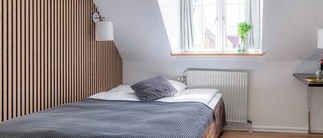 Une chambre avec un plafond en pente, une fenêtre avec des rideaux, un radiateur et une table de chevet avec une lampe.