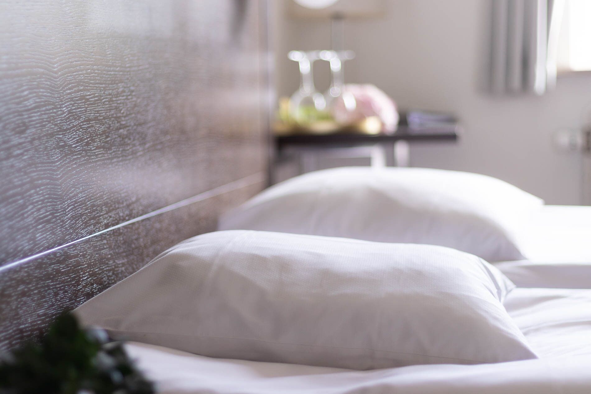 A neatly made bed with white linens and pillows, a wooden headboard, and a small table with a lamp and fruit in the background.