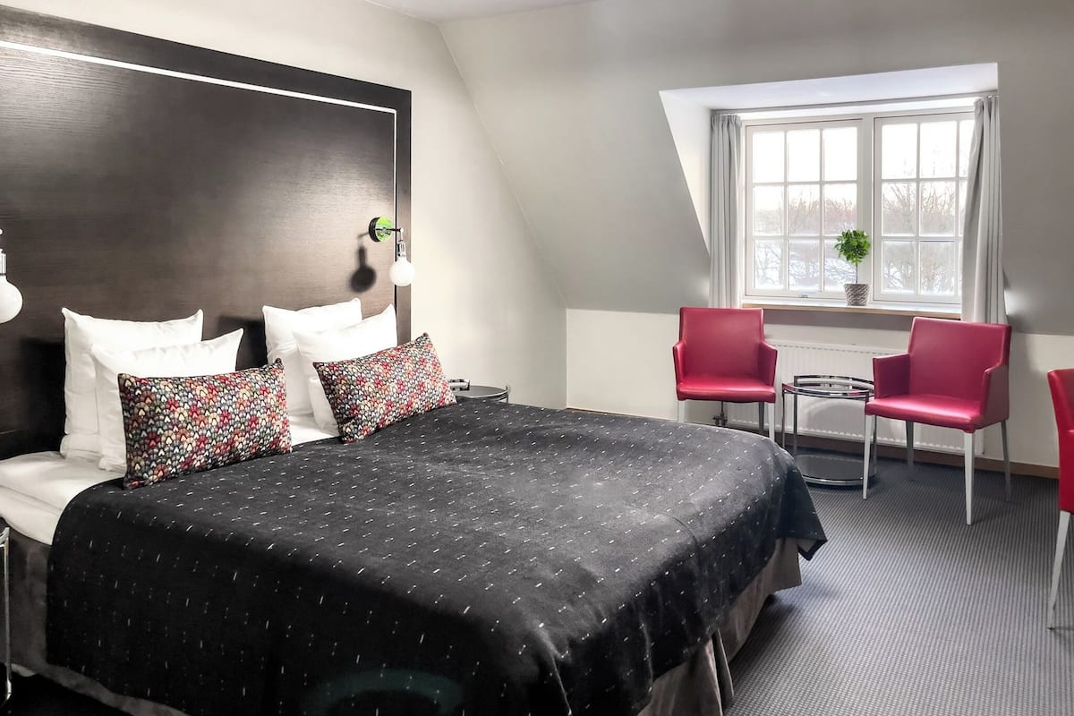 Ein modernes Hotelzimmer mit einem großen Bett, zwei roten Sesseln, einem kleinen Tisch und einem Fenster mit Blick.