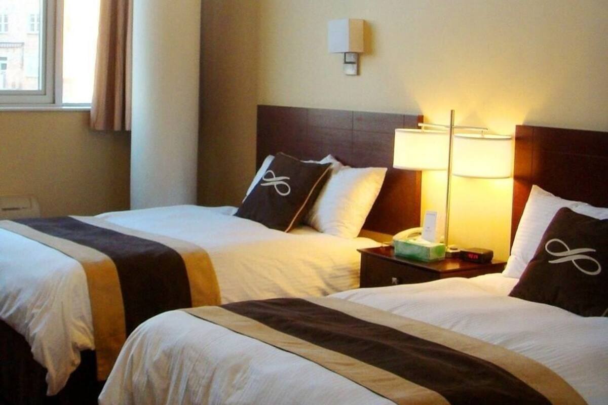 A hotel room with two beds, a window with curtains, a hanging light fixture, and a bedside table with a lamp.