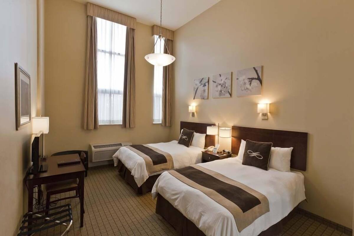 A hotel room with two beds, a desk, a chair, a lamp, and artwork on the wall.