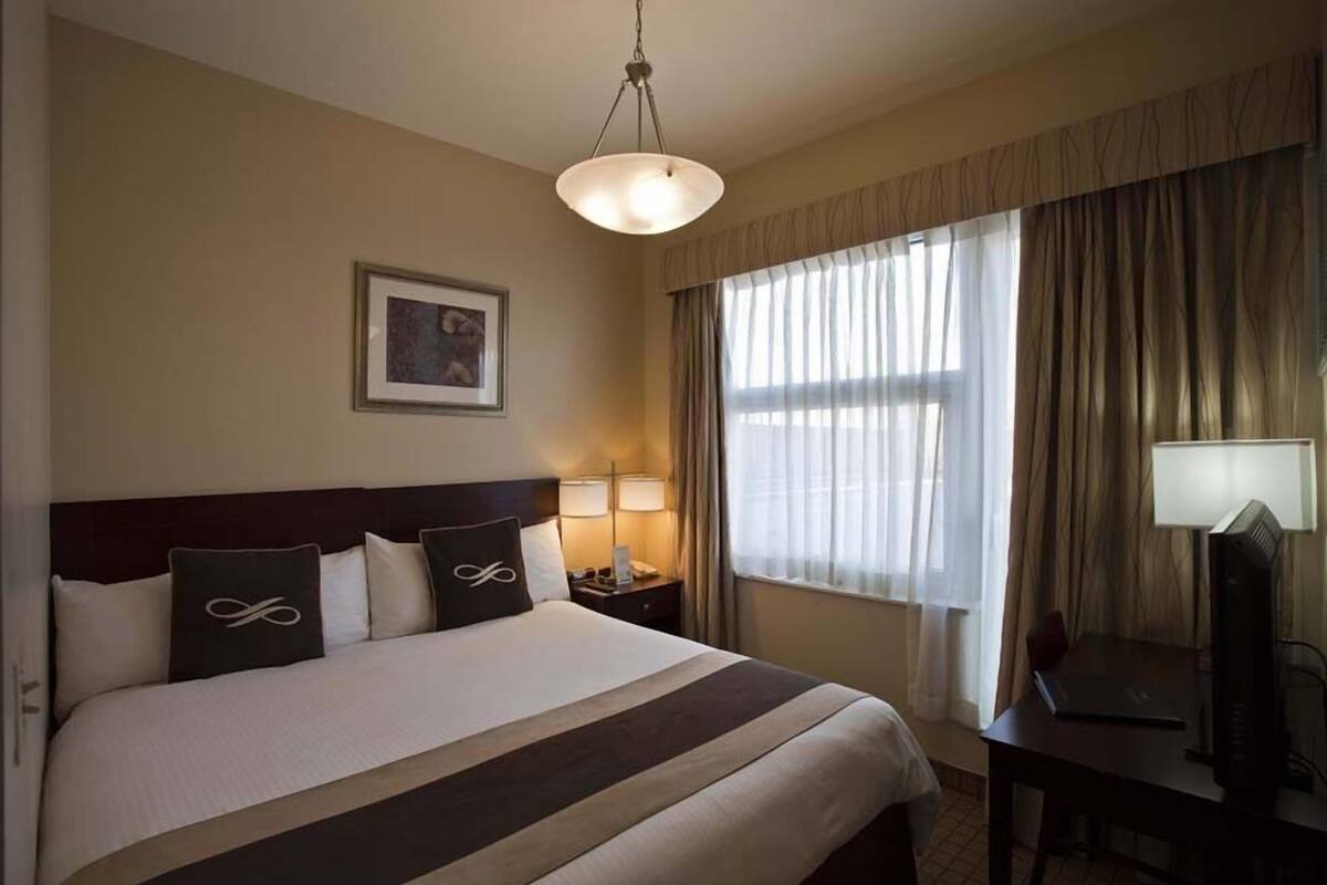 A hotel room with a bed, a desk, a lamp, a picture on the wall, and a window with curtains.