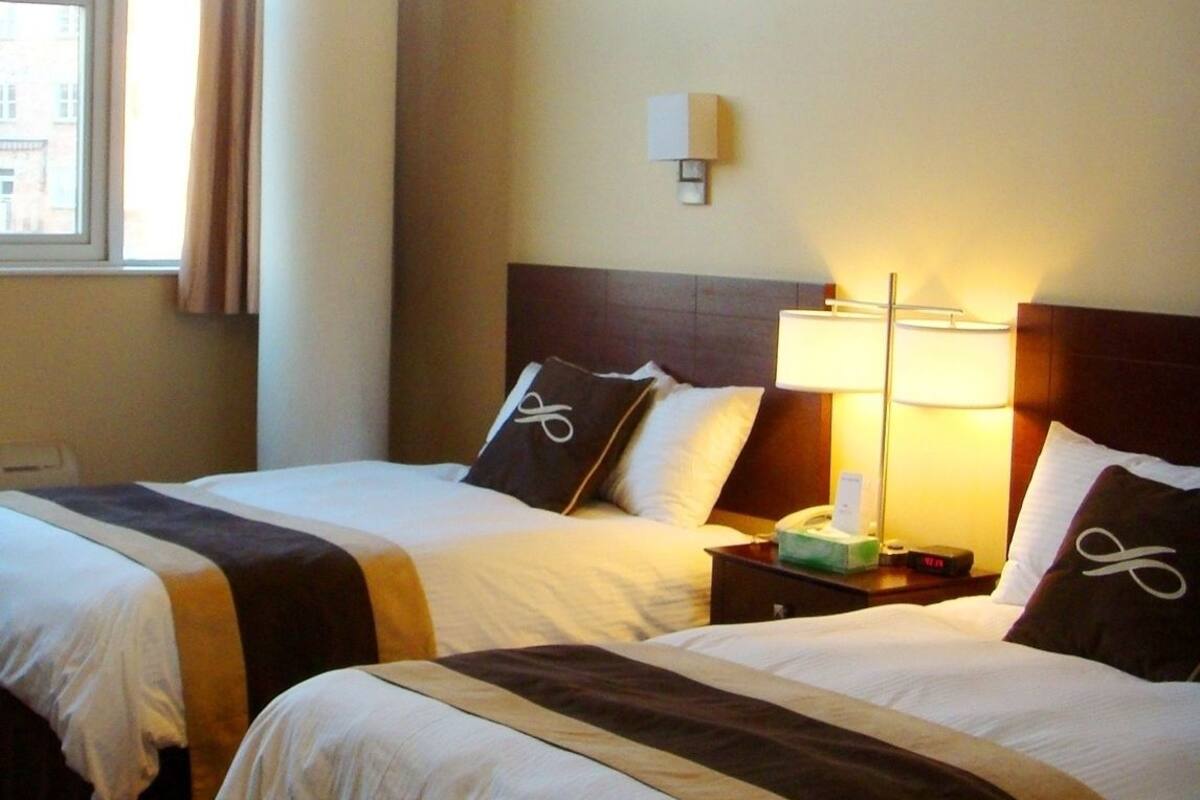 A hotel room with two beds, each with a headboard, a bedside table, a lamp, and a window with curtains.