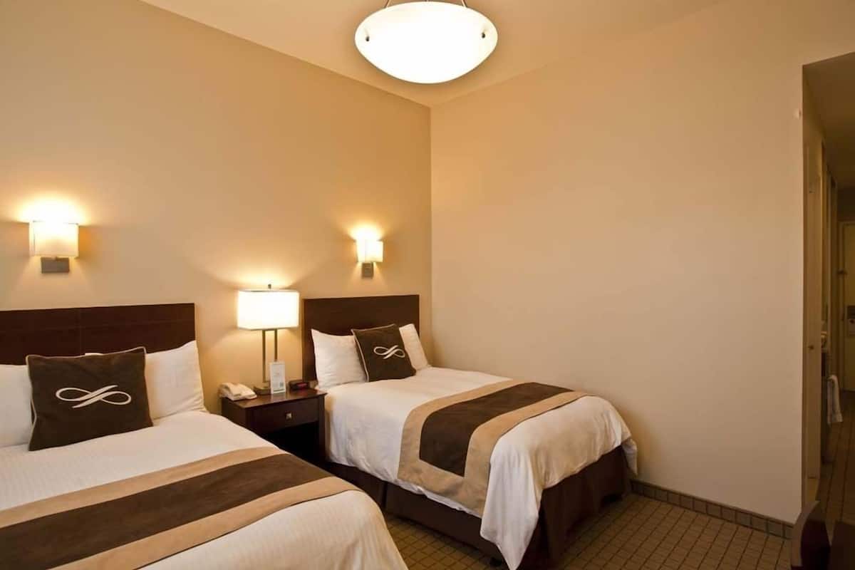 A hotel room with two beds, each with a headboard, bedside tables, and wall-mounted lamps.