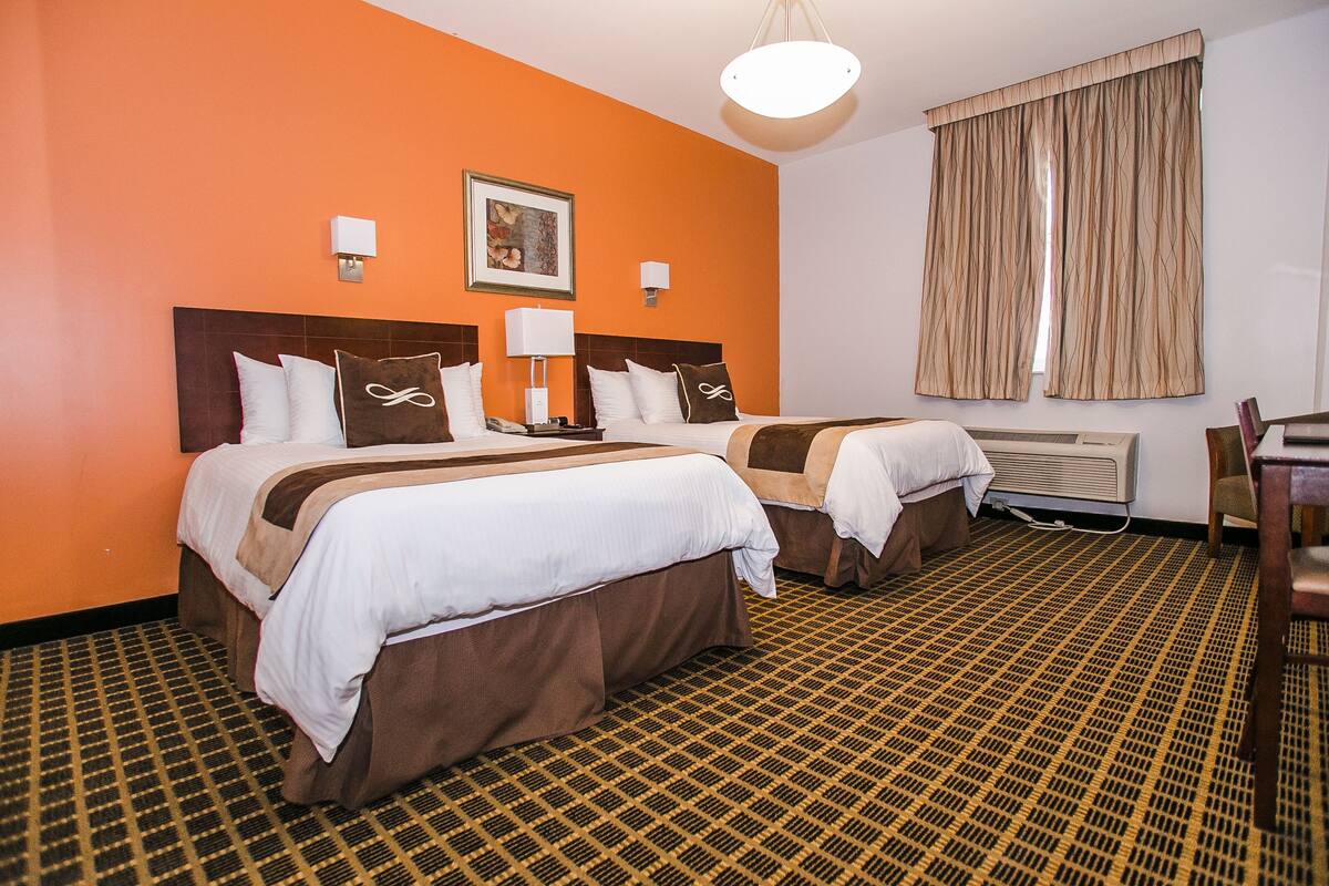 A hotel room with two beds, a large window with curtains, a patterned carpet, and a painting on the wall.