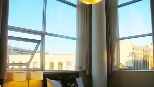 Une chambre d’hôtel avec un lit, une table de chevet dotée d’une lampe et d’un téléphone, et de grandes fenêtres offrant une vue sur les bâtiments environnants.
