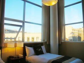 A hotel room with a bed, a nightstand with a lamp and phone, and large windows offering a view of buildings outside. - Pointe Plaza Hotel (Brooklyn)