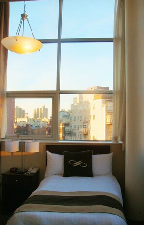 Habitación de hotel con un ventanal grande, una cama con almohada negra, una mesita de noche con lámpara y vistas a la ciudad.