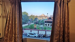A view from a window with curtains, featuring a street scene with a white car, a scooter, and a building with a red roof. - Parasol Inn Chiang Mai Old City (Chiang Mai)