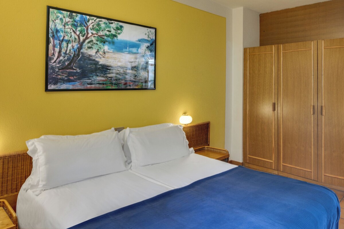 A bed with white linens, a blue blanket, and a painting of a landscape on the wall.