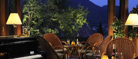 Un piano, des chaises en osier, une table avec des verres à vin, une bouteille, et une vue sur les arbres et les montagnes.