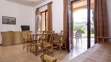 Un comedor con una mesa y sillas de madera, un televisor sobre un mueble y vistas al exterior a través de ventanas amplias.
