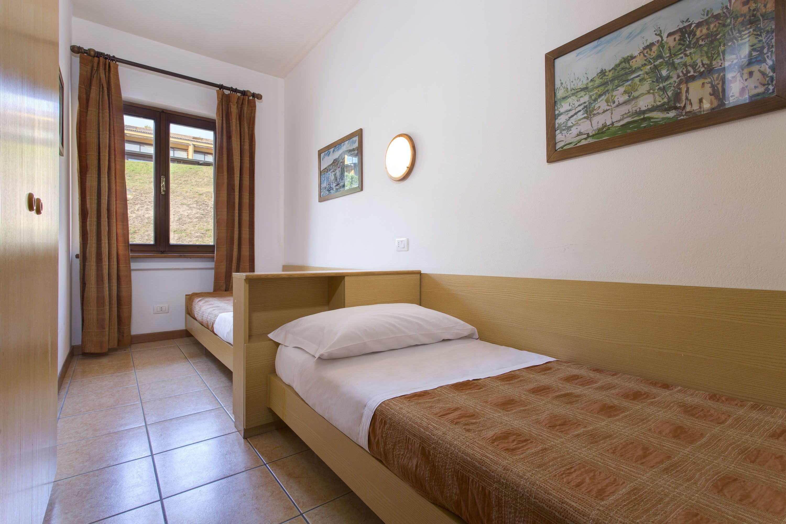 Una cama individual con cabecera de madera, una mesita de noche, una ventana con cortinas, una lámpara de pared e imagen en un marco.