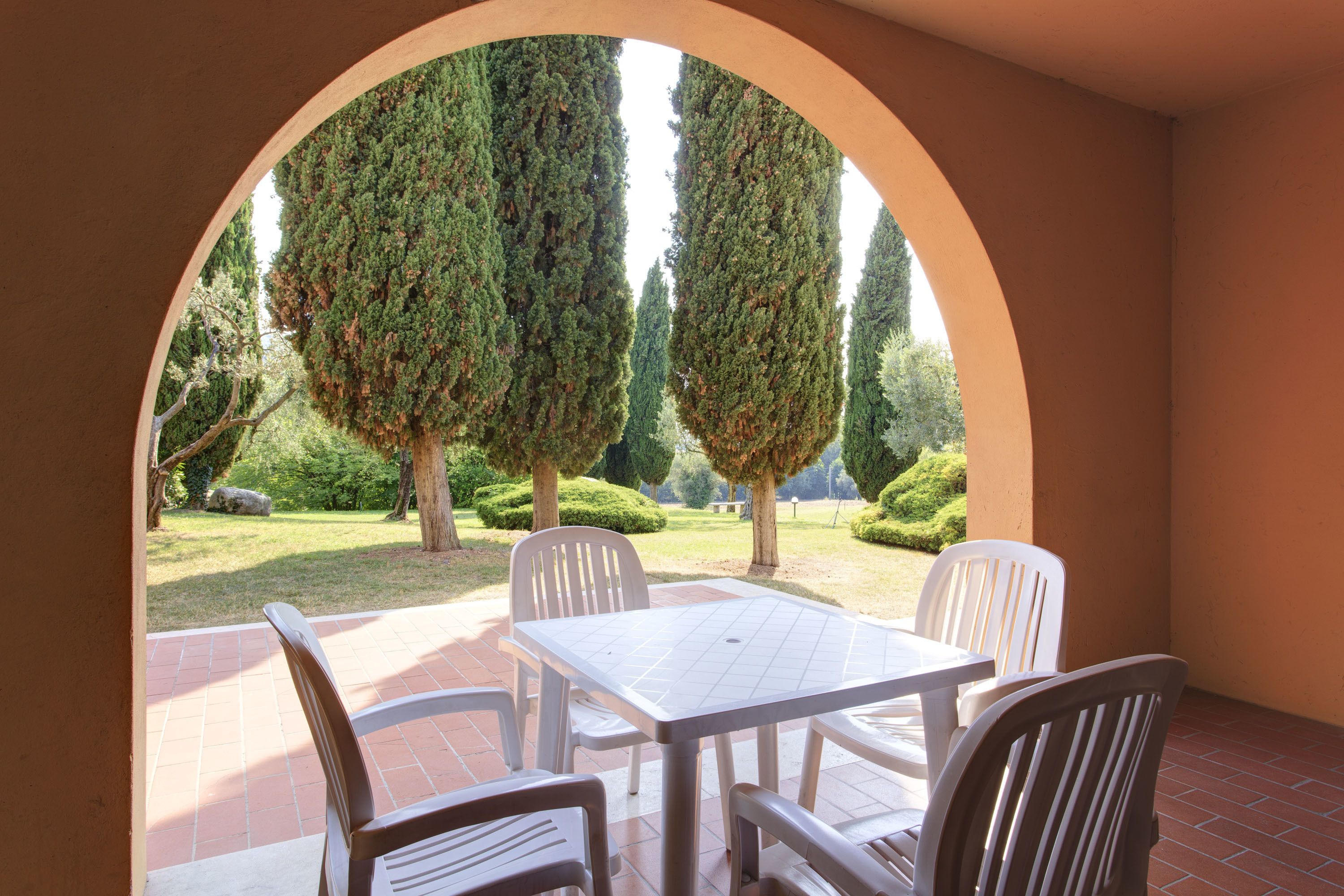 Un patio con una mesa y sillas blancas, un arco que enmarca la vista de árboles bien cuidados y un césped impecable.