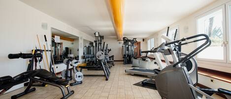 Un gimnasio con diversos equipos de ejercicio, incluyendo cintas de correr y máquinas de pesas, dispuesto en una sala amplia con ventanas grandes.