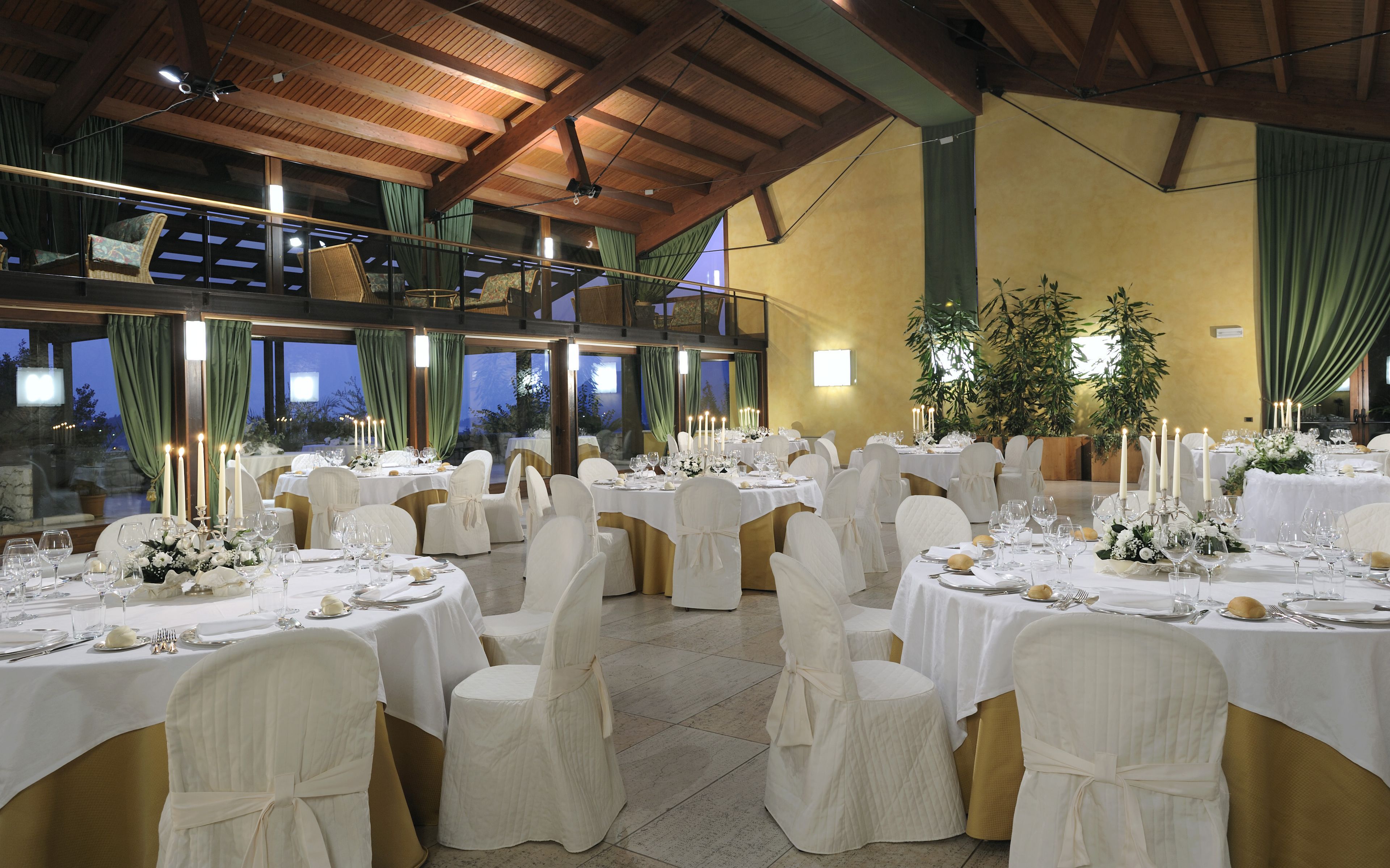 Une salle de banquet avec de nombreuses tables rondes dressées pour un événement formel, agrémentées de nappes blanches, de rubans dorés et de centres de table ornés de bougies.