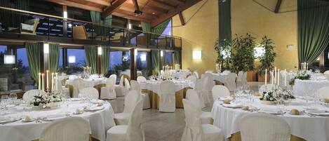 Une salle de banquet avec de nombreuses tables rondes dressées pour un événement formel, agrémentées de nappes blanches, de rubans dorés et de centres de table ornés de bougies.