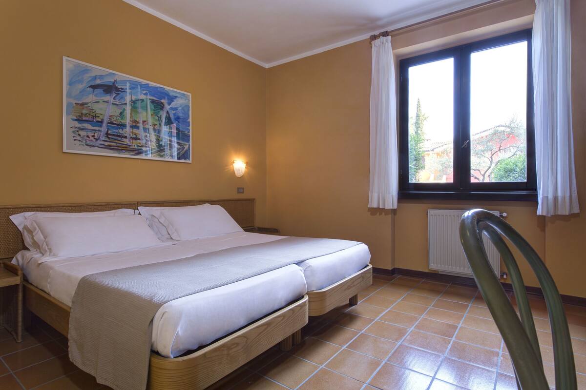 A double bed with a headboard, a painting on the wall, a window with curtains, and a radiator.