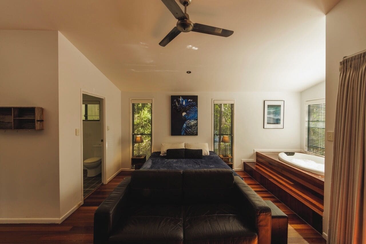 A spacious room with a large bed, a ceiling fan, and a view of a garden through the windows.