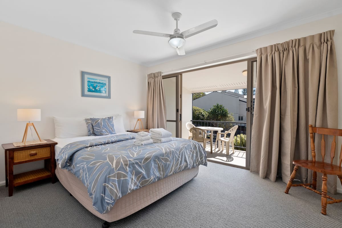 A bedroom with a bed, bedside tables, a ceiling fan, and a view of an outdoor patio with a table and chairs.